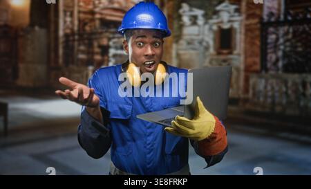 Operaio edile nero con elmetto e tute blu che tengono il computer portatile e scrivono con i guanti; concentratevi sulla produttività. Foto Stock