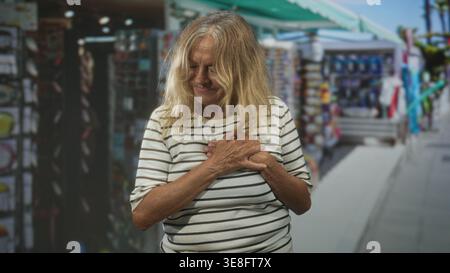 Donna anziana che stringe le mani al petto e inclina la testa all'indietro a occhi chiusi su una strada costeggiata da bancarelle del mercato; riflesso di gratitudine. Foto Stock