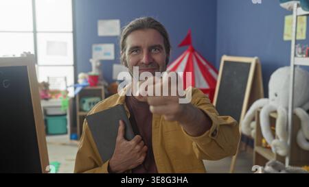 Uomo che indossa una giacca gialla che tiene il tablet sorride e punta le dita verso la fotocamera in classe; incoraggia la fiducia della connessione. Foto Stock