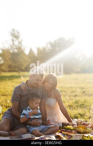 Una famiglia gioiosa trascorre del tempo insieme su una coperta, condividendo risate e cibo in un campo illuminato dal sole. Il bambino interagisce con i genitori in un bancomat caldo Foto Stock