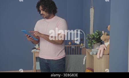 L'uomo tocca il tablet con il dito indice nel camerino mentre si avvicina per regolare il giocattolo imbottito sul comò; la cura dei genitori calma. Foto Stock
