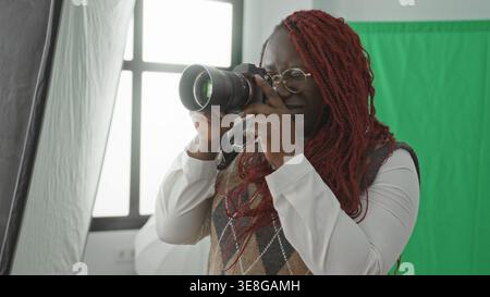 Fotografa donna con le mani in mano una fotocamera dslr, regolazione delle impostazioni in uno studio fotografico con schermo verde e illuminazione softbox; concentrazione creativa Foto Stock