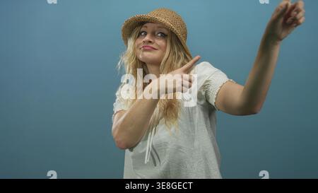 La donna punta il dito verso l'alto nello studio blu indossando un cappello di paglia e una camicia bianca, visibile a mano; direzione giocosa. Foto Stock
