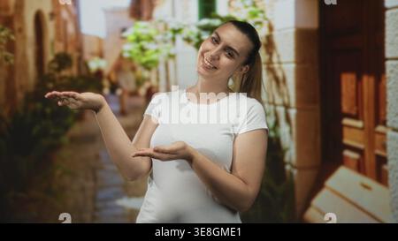 Donna che presenta le palme sorridente come se tenesse un oggetto in una stretta e soleggiata strada acciottolata incorniciata da muri in pietra e porta in legno; benvenuto Foto Stock