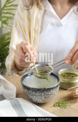 Donna setacciando polvere di matcha nel recipiente al tavolo di legno, primo piano Foto Stock