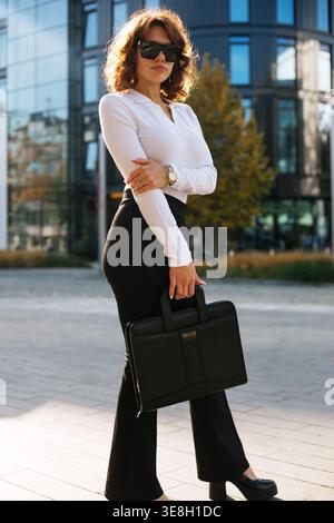 Donna in piedi in città con una borsa nera. Bella donna d'affari che indossa abiti eleganti e occhiali da sole. Look professionale aziendale. Ritratto urbano Foto Stock