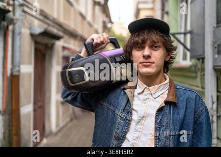 Ritratto di un giovane uomo retrò con radio portatile a spalla, stile streetwear, revival anni ottanta, cultura della musica urbana, identità giovanile sicura. Foto Stock