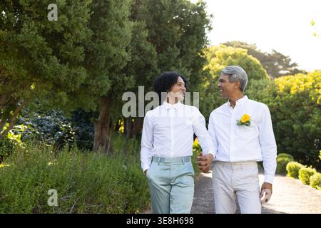 Uomini afroamericani che si tengono per mano e camminano sul sentiero del giardino, indossando camicie bianche e boutonniere Foto Stock