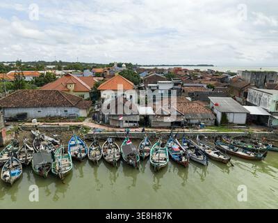 Vista aerea delle barche colorate raggruppate lungo il mercato del pesce galleggiante di Jepara, in contrasto con i toni terrosi degli edifici e del mare, Jepara, Foto Stock