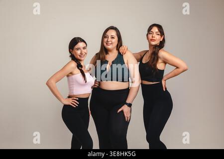 Concetto di amicizia. Bellissime e variegate amiche del fitness, tra cui una donna ricurva con fisico atletico, si mettono insieme in un abbigliamento sportivo da studio. Foto Stock
