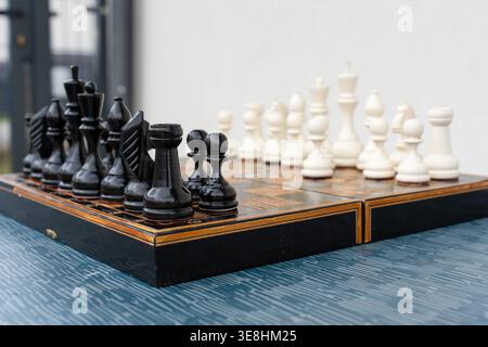 Set completo per scacchi con pezzi in bianco e nero disposti su tavola decorativa intarsiata all'aperto su un tavolo blu con sfondo ghiaia. Concetto di esterno Foto Stock