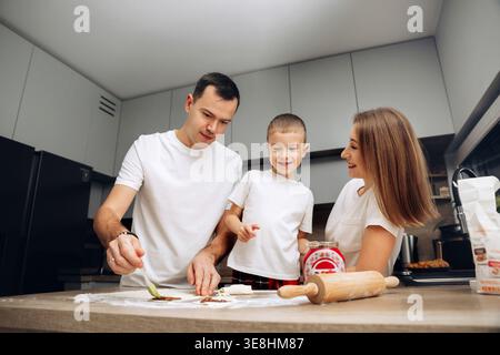 I membri della famiglia lavorano sulla cottura dei biscotti in una cucina piena di luce. Un bambino guarda e aiuta mentre gli adulti mescolano gli ingredienti, creando divertimento e divertimento Foto Stock