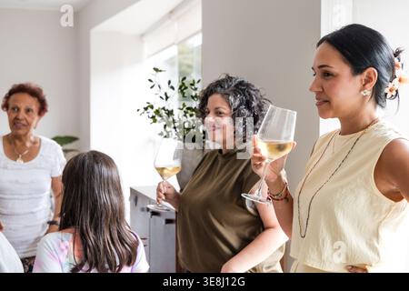 Una famiglia ispanica multigenerazionale di donne e bambini che si riuniscono in una casa luminosa e moderna. Le donne sorridono e bevono vino bianco insieme du Foto Stock