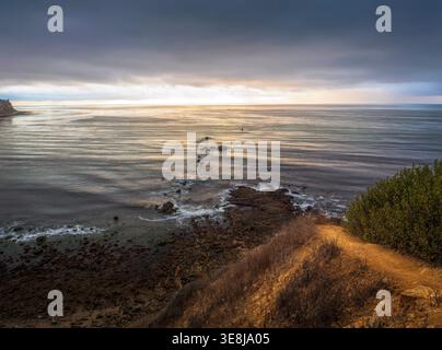 La luce dorata del tramonto sulla scogliera rocciosa costiera e l'orizzonte calmo dell'oceano con un cielo spettacolare Foto Stock