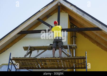 L'operaio edile si trova su un ponteggio che misura il rivestimento in legno sotto il tetto, la casa gialla, il casco di sicurezza, il progetto di miglioramento della casa, lo specialista all'aperto a. Foto Stock