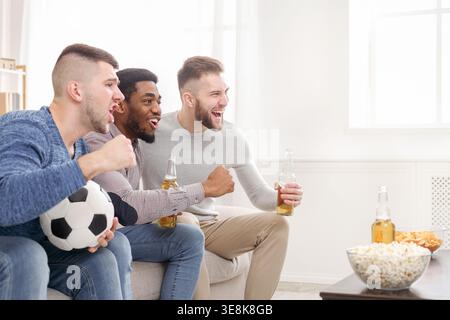 Un gruppo di amici si diverte con una partita di calcio mentre sorseggi spuntini e bevande in un accogliente soggiorno Foto Stock