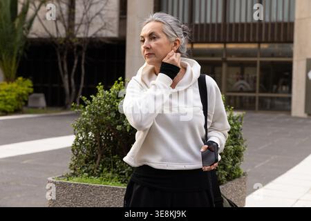 Donna anziana che fa una pausa in plaza, indossa una felpa con cappuccio bianca, tocca il collo, tiene lo smartphone vicino alla piantatrice Foto Stock