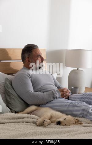 Uomo maturo seduto sul letto di casa, con pigiami grigi, che guarda a destra, cucciolo che dorme accanto alla lampada Foto Stock