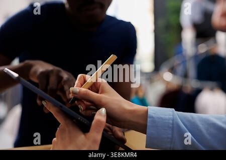 Lavoratore in possesso di un tablet, firma un rapporto online in una boutique moderna. Il dipendente del punto vendita parla con il corriere della spedizione e prepara i pacchi per la consegna nel centro commerciale. Primo piano Foto Stock