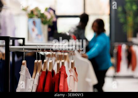 Attenzione selettiva di appendiabiti pieni di abiti alla moda in una moderna boutique, in sottofondo coppie afroamericane che fanno shopping per camicie alla moda, articoli in tessuto. Concetto di moda Foto Stock