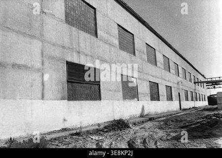 Sloviansk, 1980 anni, territorio della fabbrica AIZ. Questa foto architettonica mostra la lunga e imponente facciata di un edificio industriale in cemento con più file di finestre a griglia in vetro e pesanti feritoie di ventilazione poste sul muro. Il design solido e funzionale riflette l'estetica standard sovietica per i grandi impianti di produzione. Questa autentica fotografia d'archivio in bianco e nero conserva il patrimonio industriale, le tipiche condizioni di lavoro sovietiche e la realtà quotidiana della vita in un tranquillo Donbas prima della guerra. Foto Stock