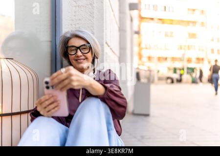 Donna anziana sorridente che si connette con lo smartphone in un ambiente urbano Foto Stock