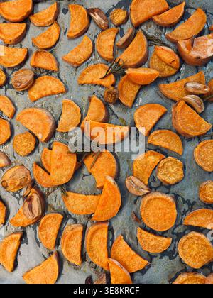Patate dolci arrostite con spicchi d'aglio e rosmarino fresco su una teglia da forno scura. Contorno vegano sano. Vista dall'alto. Cibo rustico fotogra Foto Stock