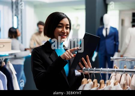 Una giovane donna asiatica sorridente che analizza le scorte di abbigliamento sul tablet digitale mentre lavora nel negozio di abbigliamento del mercato al dettaglio. Dipendente del centro commerciale che gestisce l'inventario delle merci per l'abbigliamento Foto Stock
