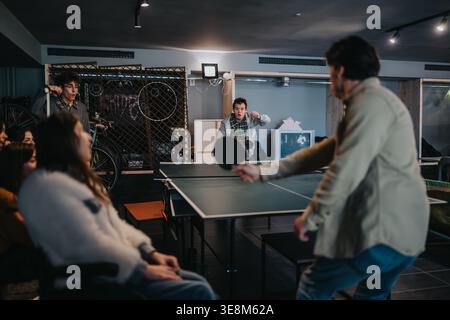 Gli studenti giocano a ping pong in una sala comune del college con gli amici che guardano e reagiscono Foto Stock