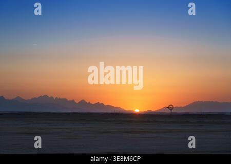 Il tramonto proietta un caldo bagliore su un vecchio mulino a vento che si erge graziosamente nel deserto, un simbolo senza tempo della tranquillità rurale Foto Stock