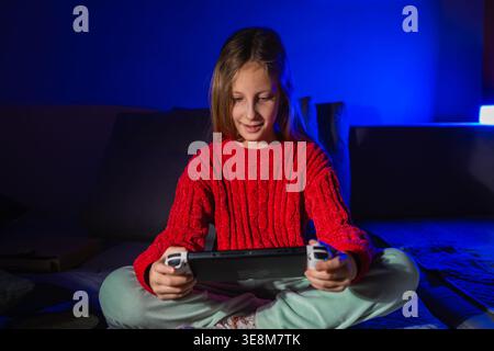 Ragazza sorridente che si diverte con un videogioco portatile mentre si siede in una stanza buia con luce blu Foto Stock