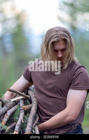 Giovane con lunghi capelli biondi che indossa una T-shirt marrone e pantaloncini neri, tenendo un fascio di rami tagliati tra le braccia. Foto Stock
