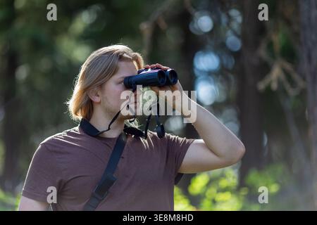 Foto con profilo laterale di un giovane con lunghi capelli biondi che guarda attraverso un binocolo in un ambiente forestale. Foto Stock