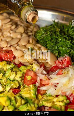 Uno scatto verticale in primo piano che cattura il momento in cui l'olio d'oliva viene versato da una bottiglia di vetro su una ciotola in acciaio inox riempita con ingredienti freschi per insalata Foto Stock