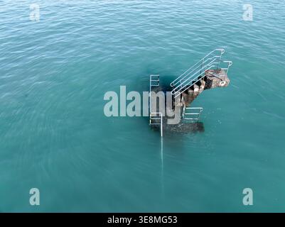 Vista aerea di gradini in pietra con ringhiere metalliche che scendono nel mare turchese, creando un netto contrasto tra la struttura artificiale e. Foto Stock