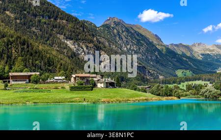 Una vista pittoresca di case in pietra annidate in prati verdi accanto alle vibranti acque turchesi del Lago di Brusson, adagiate contro le montagne in Italia Foto Stock