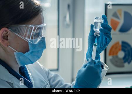 Medico o infermiere che indossa guanti blu e camice da laboratorio riempie la siringa dal flaconcino di vetro, concentrandosi sul processo di vaccinazione. Moderno concetto di assistenza sanitaria e immunizzazione in un ambiente ospedaliero luminoso Foto Stock