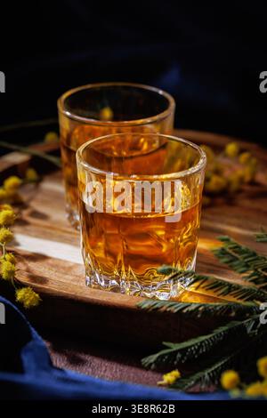 Due bicchieri di whisky o brandy su vassoio di legno con Mimosa Flowers Spring Alcohol Concept Foto Stock