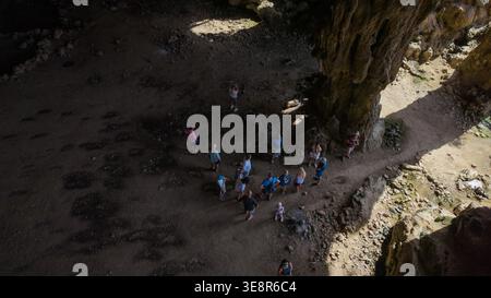 Vista aerea dei turisti che si trovano all'interno di una grande grotta rocciosa sul mare con luce del sole che crea forti ombre sul pavimento sabbioso e sulle pareti della scogliera. Gradi Foto Stock