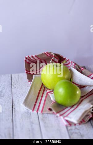 Due mele verdi poggiano su un panno a righe rosso e beige collocato su un tavolo di legno chiaro in cucina. La luce naturale illumina la scena. Foto Stock