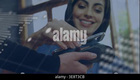 Donna ispanica sorridente che indossa un foulard scuro e la parte superiore blu che tiene il telefono sul terminale POS del bar Foto Stock
