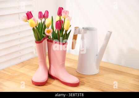 Bellissimi fiori di tulipani con stivali in gomma rosa e annaffiatoio su un tavolo di legno vicino a pareti bianche interne Foto Stock
