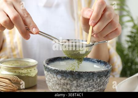Donna setacciando polvere di matcha nella ciotola a tavola, primo piano Foto Stock