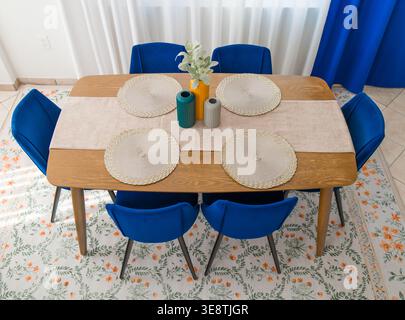 La foto ad alto angolo di una sala da pranzo domestica mostra un tavolo in legno con sedie in velluto blu e un tappeto colorato con motivi floreali sul pavimento. Foto Stock
