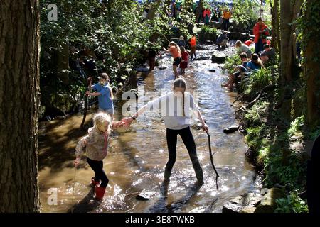 Due ragazze che attraversano il fiume Porter tenendosi per mano con altri partecipanti alle retrovie durante la gara di anatra pasquale all'Endcliffe Park, Sheffield, Regno Unito, il 6 aprile Foto Stock