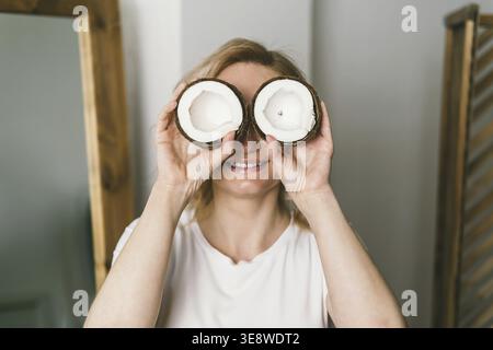 Una bella ragazza tiene in mano metà di una noce di cocco e si copre gli occhi con loro. Donna che tiene in mano una noce di cocco fresca Foto Stock