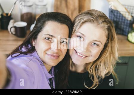 Le amiche ridono allegramente e scattano selfie in cucina. Due ragazze sono fotografate al telefono. Bruna e bionda si divertono. Donne che guardano sma Foto Stock