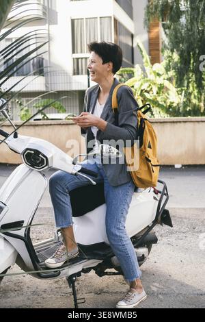 Una donna di mezza età prende il suo scooter bianco. Donna d'affari con zaino e telefono in mano sorride allegramente. Donna con taglio di capelli corto e buio Foto Stock