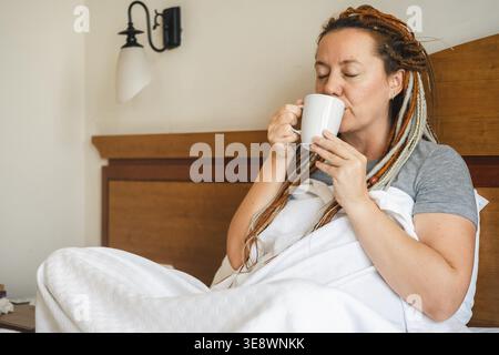 Donna di mezza età che beve caffè mattutino a letto. La donna con i dreadlocks sulla testa sorride contentosamente Foto Stock