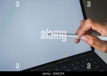 Un primo piano ad angolo alto che mostra la mano di una persona che tiene in mano uno stilo digitale bianco e lo punta verso uno schermo bianco luminoso del tablet. Foto Stock